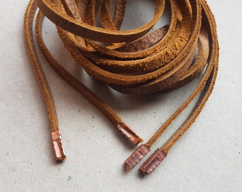 Puede incluir: Cordones de cuero marr&oacute;n enrollados con terminales de color cobre. Los cordones tienen un tono c&aacute;lido y terroso, y los terminales son rectangulares. El cuero parece texturizado y desgastado, lo que sugiere una est&eacute;tica vintage o r&uacute;stica.