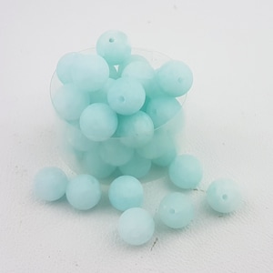 May include: A close-up of a container filled with light blue silicone beads. The beads are round and have a matte finish. There are also several beads scattered on a white surface.
