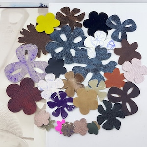 Op de afbeelding: Een verzameling leren bloemuitsparingen in tinten bruin, geel, paars, roze en zwart. Sommige bloemen hebben een textuur, andere zijn glad. De uitsparingen zijn gerangschikt op een wit oppervlak.