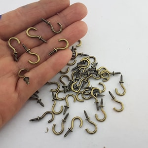 Può includere: Una collezione di piccoli ganci a tazza in metallo color ottone antico, ciascuno dotato di una base a vite filettata e una parte superiore curva. Questi elementi di fissaggio sono progettati per essere montati su superfici per appendere.