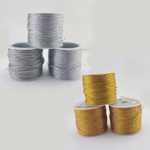 May include: Three spools of metallic thread, two gold and one silver, are wound on white plastic spools. The spools are arranged in a pyramid shape.
