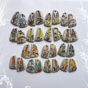 Op de afbeelding: Een verzameling beige keramische hangers, elk met de hand beschilderd met kleurrijke bloemmotieven. De hangers zijn in paren en tonen verschillende bloemen in tinten blauw, geel, rood en paars. Elke hanger heeft een klein gaatje aan de bovenkant om op te hangen.