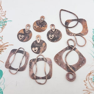 May include: A selection of copper-coloured metal pendants and charms. The pendants display diverse shapes, including circles, squares, and teardrops, with heart cutouts and loop attachments. The metal has a rustic, mottled appearance.