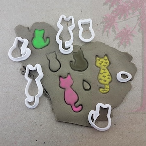 May include: A collection of cat-shaped cookie cutters and cat-shaped impressions in clay. The clay has green, pink, and yellow cat shapes. The cutters are white plastic. The image is a top-down view.