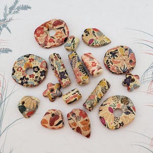 Puede incluir: Una colección de cuentas de cerámica variadas en diferentes formas, incluyendo anillos, cilindros, estrellas y formas irregulares. Cada cuenta presenta una base de color crema con un patrón floral y botánico colorido en tonos rojos, azules y amarillos.