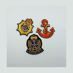 Könnte beinhalten: Drei gestickte Patches mit goldenen und roten Fäden. Der erste Patch ist ein Kreis mit einem goldenen Anker und dem Text "US CHAMPION". Der zweite Patch ist ein roter Anker mit goldenen Fäden. Der dritte Patch ist ein Kreis mit einem goldenen Anker und dem Text "COUNTRY".
