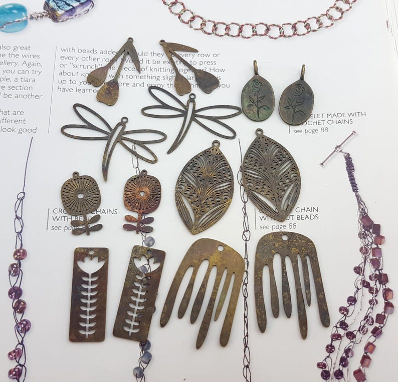 May include: A collection of bronze-colored metal jewelry findings, including dragonfly, leaf, and floral shapes. Some pieces have intricate cut-out designs. The findings are arranged on a printed page with text.