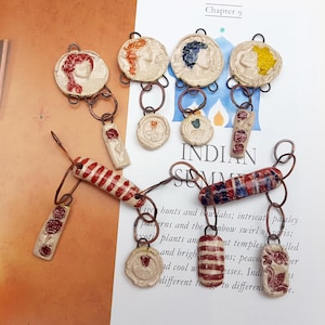 May include: A selection of handcrafted ceramic pendants and charms in diverse forms. The pendants showcase relief designs and are linked with copper-toned metal rings. The colour scheme includes red, blue, yellow, and white.