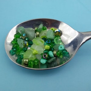 May include: A silver spoon filled with green and clear glass beads. The beads are various shapes and sizes, including round, oval, and square.