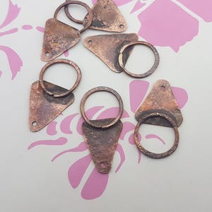 Puede incluir: Primer plano de varios hallazgos metálicos de color cobre. Cada hallazgo tiene forma triangular con un pequeño agujero en el extremo puntiagudo y un anillo circular adjunto. El metal tiene un aspecto rústico y envejecido.