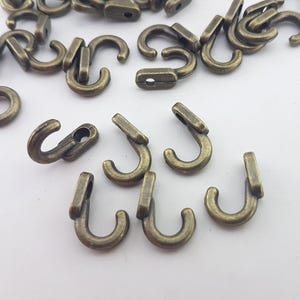 Può includere: Una collezione di piccoli ganci in metallo color bronzo antico. Ogni gancio presenta un design curvo a forma di J con una base rettangolare piatta contenente un singolo foro circolare per il fissaggio. La finitura ha un aspetto metallico.