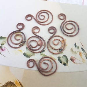 Könnte beinhalten: Sieben Kupferspiralen mit einer einzelnen Schlaufe an jeder Seite. Die Spiralen sind alle unterschiedlich groß und in einem zufälligen Muster angeordnet.