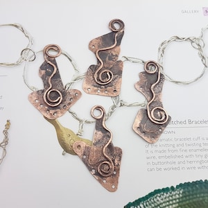 Puede incluir: Cuatro colgantes de metal de color cobre con una superficie texturizada y un bucle de alambre. Cada colgante tiene una forma y un diseño únicos.