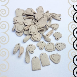 Könnte beinhalten: Eine Sammlung kleiner, beigefarbener Keramikanhänger und -anhänger in verschiedenen Formen, darunter Herzen, Anhänger und abstrakte Designs. Einige Teile haben strukturierte Oberflächen und kleine Löcher zum Auffädeln. Die Artikel sind auf einer weißen Oberfläche verstreut.