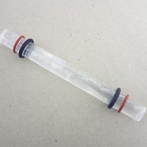 May include: A clear, cylindrical glass object with a red and black ring on each end. The object appears to be a glass rod or tube, possibly for scientific or laboratory use. The rings are made of rubber or a similar material.