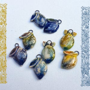 Op de afbeelding: Een verzameling handgemaakte keramische hangers in verschillende kleuren, waaronder blauw, groen en geel. Elke hanger heeft een unieke vorm met een kleine lus om op te hangen. De hangers zijn gerangschikt op een witte stoffen achtergrond.