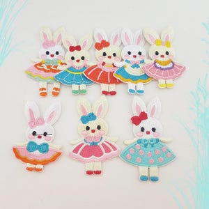 Op de afbeelding: Geborduurde patches met witte konijnen in roze, blauwe, oranje en rode jurken. Elk konijn heeft een strik en draagt een jurk met een bijpassende strik. De patches zijn ontworpen in cartoonstijl.