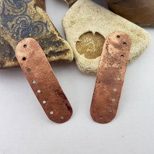 May include: Two copper-coloured metal pieces with rounded edges and multiple holes. The pieces are on a white surface.
