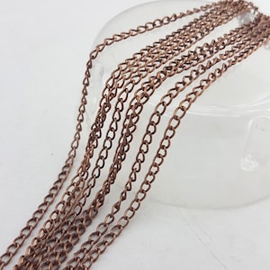 Puede incluir: Primer plano de varias hebras de cadena de color cobre. Las cadenas están hechas de pequeños eslabones ovalados y están dispuestas en líneas paralelas. Las cadenas descansan sobre una superficie clara y redondeada, con un fondo blanco.