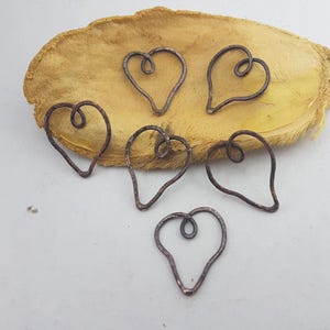 May include: Six handcrafted copper heart-shaped charms with a rustic, hammered texture. Each heart features a looped design at the top. The charms are displayed on a textured, tan-colored surface, creating a warm, earthy aesthetic.