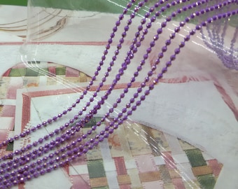 May include: A close-up of a purple chain with a metallic sheen. The chain is made of small, round beads and is lying on a white surface.