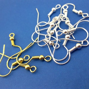 May include: A close-up of a pile of silver and gold earring hooks. The hooks are made of metal and have a small ball at the end. The hooks are on a blue background.