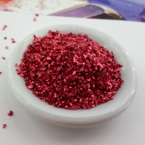 Könnte beinhalten: Eine kleine weiße Schale gefüllt mit rotem Glitter. Der Glitter besteht aus kleinen, unregelmäßigen Metallstücken.