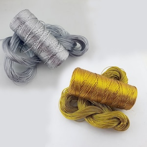 Könnte beinhalten: Zwei Spulen metallischen Fadens, eine silberne und eine goldene. Der silberne Faden ist kompakt gewickelt, mit losen Strähnen. Der goldene Faden ist ebenfalls gewickelt und hat ein glänzendes Aussehen. Geeignet für Bastelarbeiten.