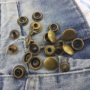 Può includere: Primo piano di un gruppo di bottoni a pressione in metallo color oro antico su uno sfondo di denim blu.