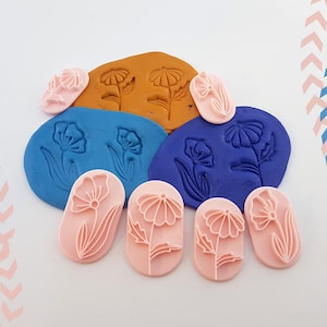 May include: A selection of pink flower-shaped stamps and imprints in blue and brown clay. The stamps feature detailed floral designs, including petals and stems. The image showcases the stamps and their imprints, highlighting their use in crafting and art projects.