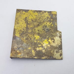 May include: A rectangular piece of metal with a cut-out section on one side. The metal has a mottled appearance with yellow and brown hues, suggesting age or weathering. The surface is textured.
