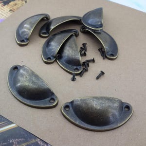 Peut inclure: Ensemble de poignées cuvette de couleur bronze antique et de petites vis noires. Les poignées, en forme de demi-lune incurvée, comportent deux trous de fixation. Idéales pour la restauration de meubles ou les projets de bricolage.