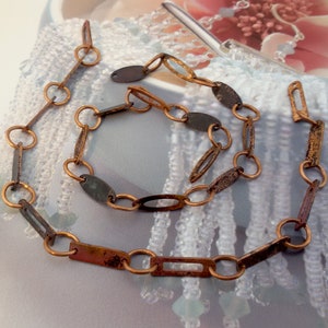 Könnte beinhalten: Ein Halsketten- und Armband-Set mit einem rustikalen, antiken Design. Der Schmuck besteht aus kupferfarbenen Metallgliedern in verschiedenen Formen, darunter Kreise, Ovale und Rechtecke. Das Metall hat ein verwittertes, gealtertes Aussehen mit dunklen Akzenten.