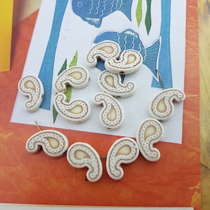 May include: Ivory-colored paisley-shaped beads with a light brown paisley design. The beads are scattered on a colorful background with a fish illustration. These beads are ideal for jewelry making and craft projects.