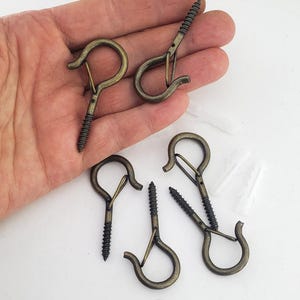 May include: A set of antique brass-finished metal screw hooks with integrated spring-loaded safety latches. Each hook features a threaded screw base for mounting and a circular loop design. Three white plastic wall anchors are included in the set.