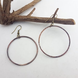 May include: A pair of circular hoop earrings crafted from hammered copper with a darkened, weathered patina. Each earring features a thin, round metal frame suspended from a simple brass-toned hook wire, showcasing a textured surface.