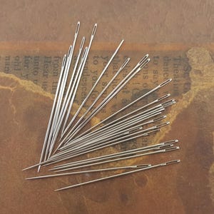 May include: A selection of silver sewing needles scattered on a brown surface. The needles are slender, with a small eye at one end and a sharp point. The background has faded text.