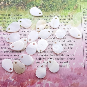 May include: A collection of 15 white mother of pearl shell teardrop shaped pendants with a hole at the top. The pendants are scattered on a pink and white patterned surface.