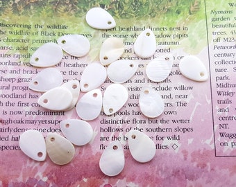 Puede incluir: Una colección de 15 colgantes en forma de lágrima de nácar blanco con un agujero en la parte superior. Los colgantes están esparcidos sobre una superficie con estampado rosa y blanco.