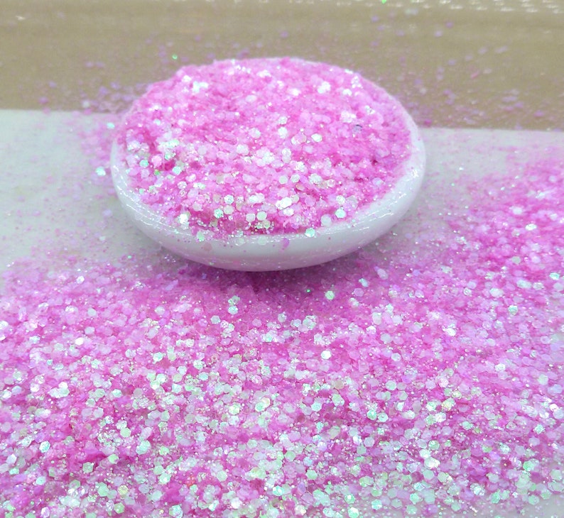 以下が含まれることがあります： 小さな白いボウルに入ったピンクと白の虹色のグリッター。グリッターはばらばらで、さまざまなサイズがあります。