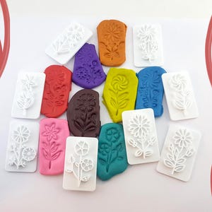 May include: A selection of colourful clay impressions and white rectangular flower-shaped stamps. The clay is in various colours, including orange, purple, red, brown, yellow, blue, pink, and green. The stamps have detailed floral designs.