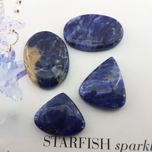 Op de afbeelding: Vier gepolijste blauwe en witte sodaliet cabochons in verschillende vormen, waaronder ovaal, driehoekig en druppelvormig.