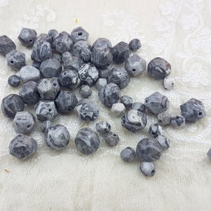 Pode incluir: Uma coleção de contas de pedras preciosas cinzentas e brancas facetadas. As contas variam em tamanho e forma, com uma mistura de formas geométricas e arredondadas. As pedras têm uma aparência marmoreada, com padrões de redemoinho de cinza e branco. Adequado para fazer joias.