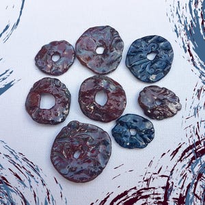 May include: A collection of eight irregularly shaped, glazed ceramic beads with a hole in the center. The beads are primarily dark red and blue, with a glossy finish. The beads are arranged on a white surface.