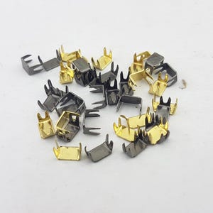 Puede incluir: Una colección de pequeños remaches metálicos en forma de U, en oro y gris plomo. Cada remache tiene cuatro púas que se extienden desde la parte superior. Los remaches están dispersos sobre una superficie blanca, mostrando su brillo metálico y diseño detallado.