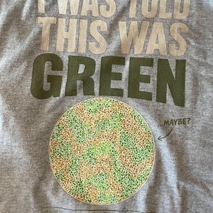 K&ouml;nnte beinhalten: Ein graumeliertes T-Shirt mit dem Text I WAS TOLD THIS WAS GREEN in gro&szlig;en Buchstaben. Darunter befindet sich ein kreisf&ouml;rmiges Farbsehtest-Muster aus gr&uuml;nen und beigen Punkten mit dem Text ...MAYBE? und einem Feld mit der Aufschrift COLOR ID: UNKNOWN.