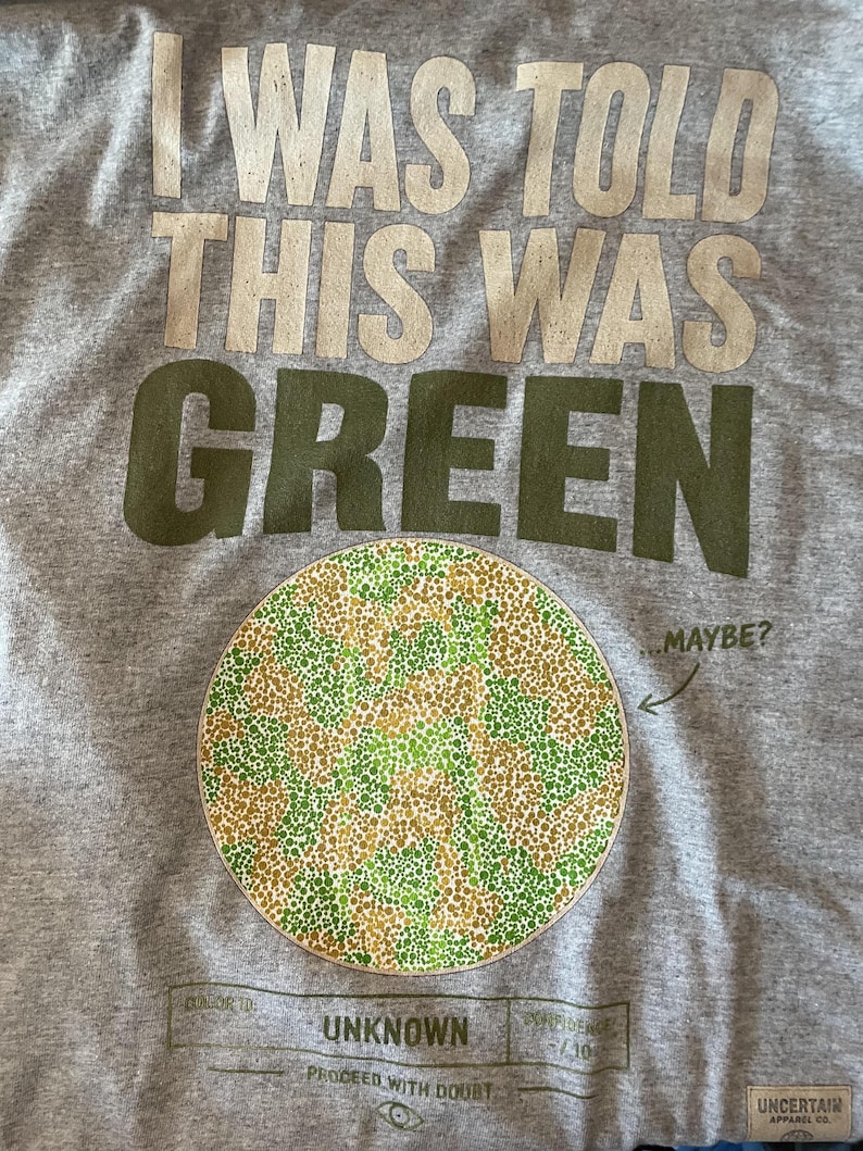 K&ouml;nnte beinhalten: Ein graumeliertes T-Shirt mit dem Text I WAS TOLD THIS WAS GREEN in gro&szlig;en Buchstaben. Darunter befindet sich ein kreisf&ouml;rmiges Farbsehtest-Muster aus gr&uuml;nen und beigen Punkten mit dem Text ...MAYBE? und einem Feld mit der Aufschrift COLOR ID: UNKNOWN.