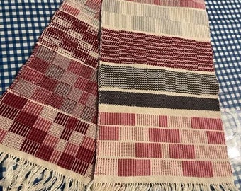 May include: A woven table runner with a red, white, and gray geometric pattern. The runner has a fringe on both ends.