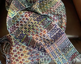 May include: A colorful woven scarf with a geometric pattern and fringe. The scarf is made of a soft, textured fabric and is folded on a brown leather chair.