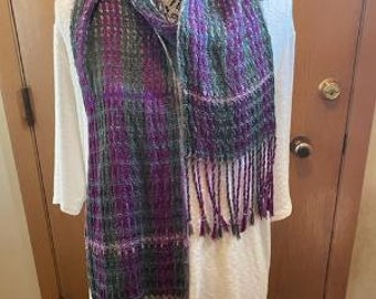 May include: A purple, green, and white woven scarf with fringe. The scarf is draped over a white shirt.